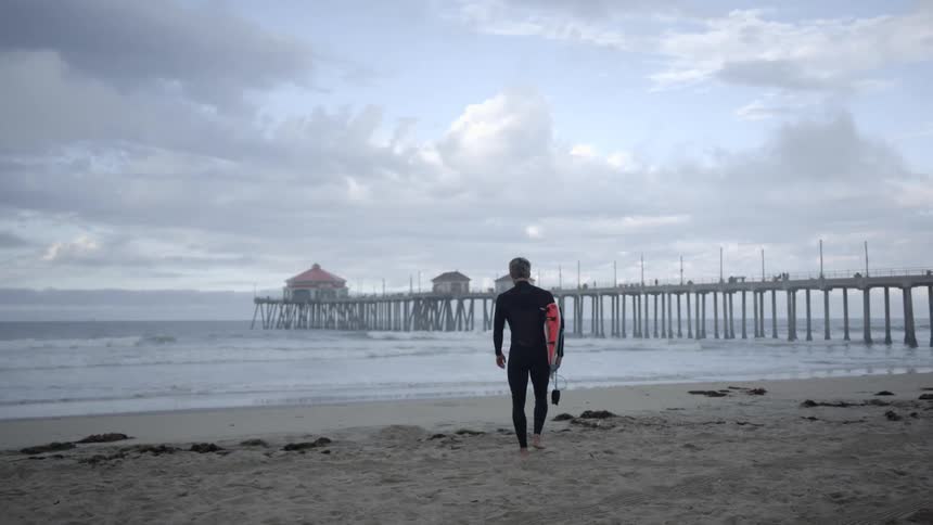 A man carrying a surfboard walks across the beach towards the water