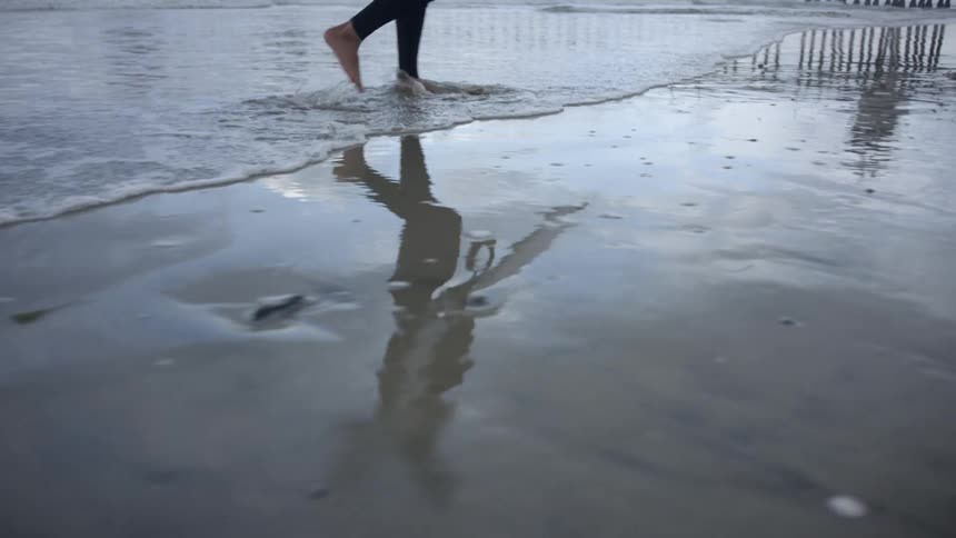Young man wearing a wet suit carries a surfboard while walking on the wet beach sand
