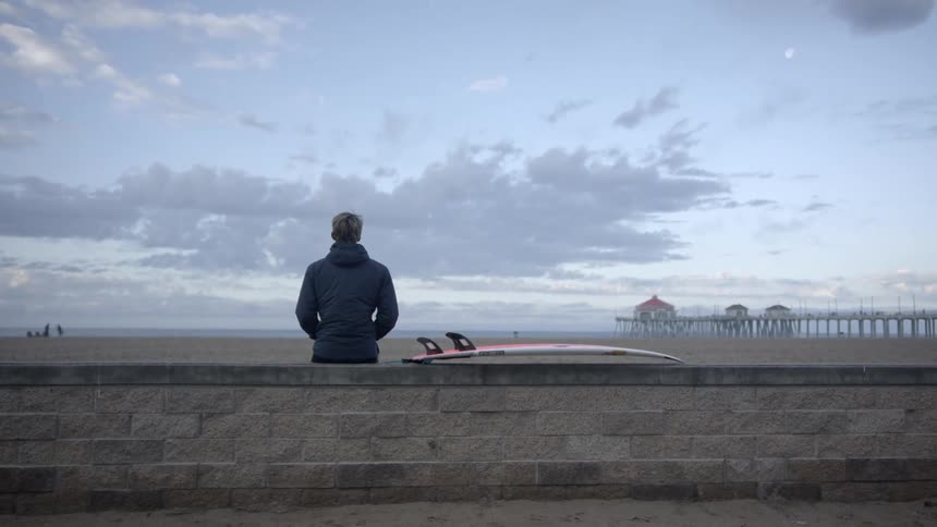 Man stands on beach leaning on wall looking at the ocean with a pier in the distance on one side and people playing on the beach on the other side
