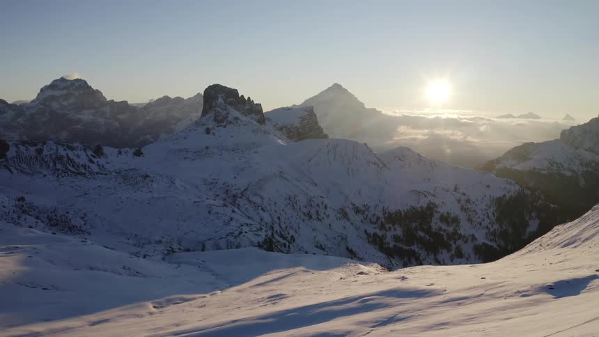 An aerial view of snowy mountains with a blue sky and the sun low in the sky