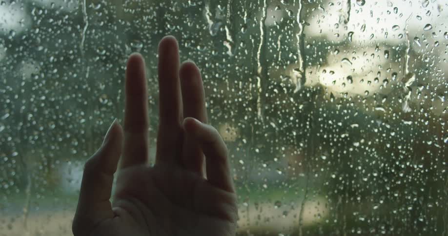 A hand is against the interior of a window touching the glass as rain falls outside and water streams down the exterior glass