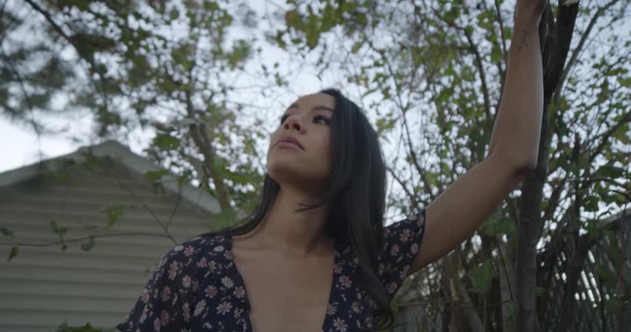 A young woman holds onto the thin trunk of a small tree as she gazes up at the sky in the backyard of a house during the day