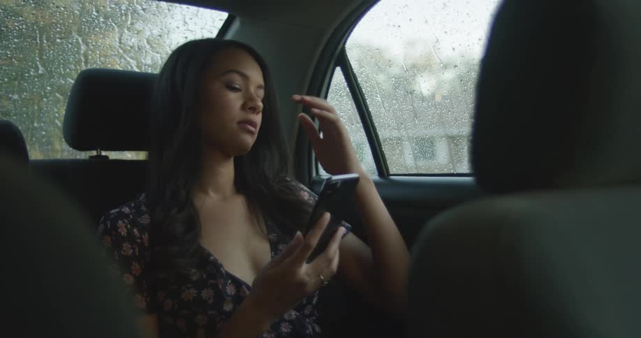 A woman sits in the back seat of a vehicle looking at her phone with a depressed expression