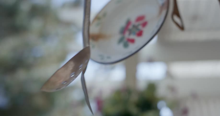Mobile made of dishes and utensils hangs outdoors under garden structure