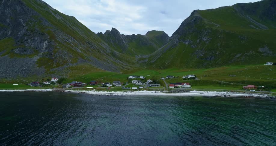 Small town with a few houses behind the sea just past a small white sand beach all of this is at the bottom of a grassy mountain range