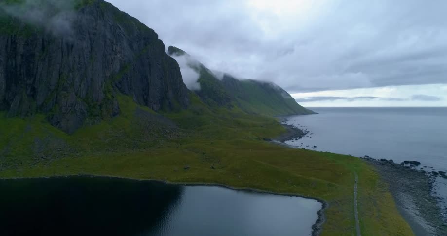A rocky shoreline at the bottom of a mountain with a grassy area before a smaller pond or lake at sunrise or sunset on a foggy day