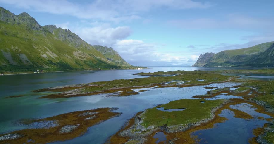 Car drives past scattered buildings hugging foot of mountain and bordered by lake