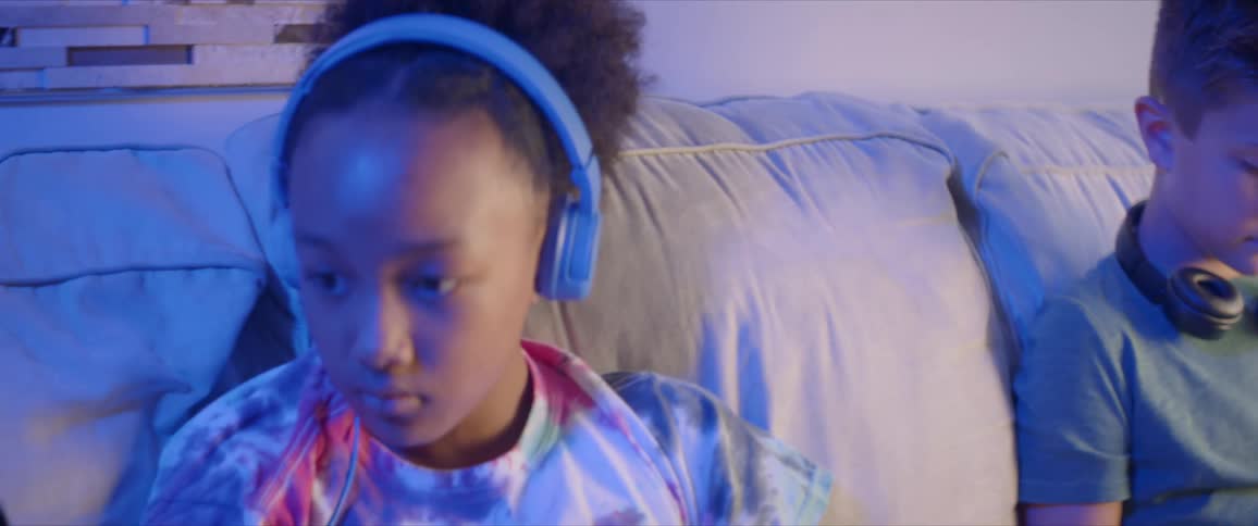 Two children sit on a couch one with headphones off and around his neck one with her head phones on and looking at two devices
