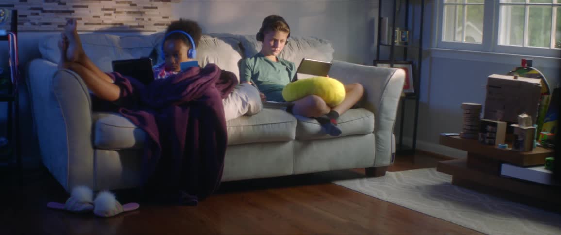 Girl wearing blue headphones and boy with black headphones and a green shirt sit on sofa and uses tablet and hand calculator in the living room at home