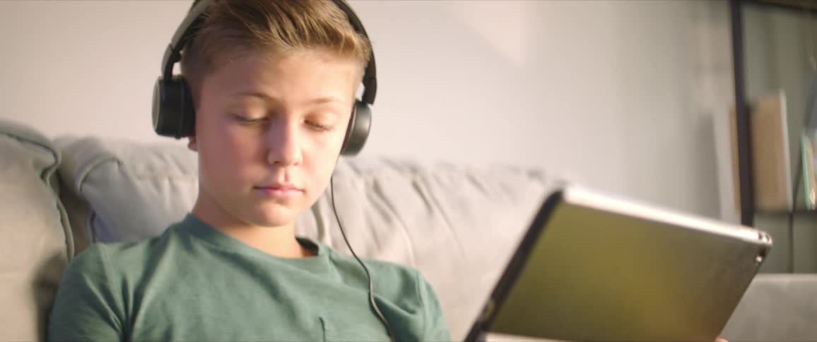 A bog sits on a couch wearing headphones and holding a tablet in his lap writes on a piece of paper