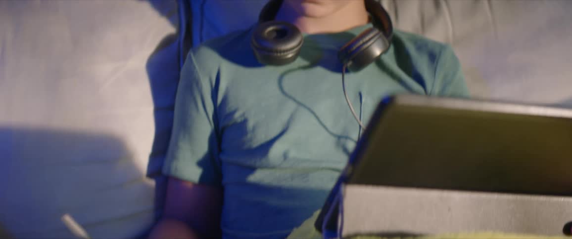 A boy sitting on a sofa in a house writes in a notebook while glancing at the screen of a computer tablet