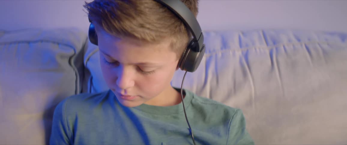 A boy sits on a couch with a laptop and wearing headphones writing on a piece of paper beside him