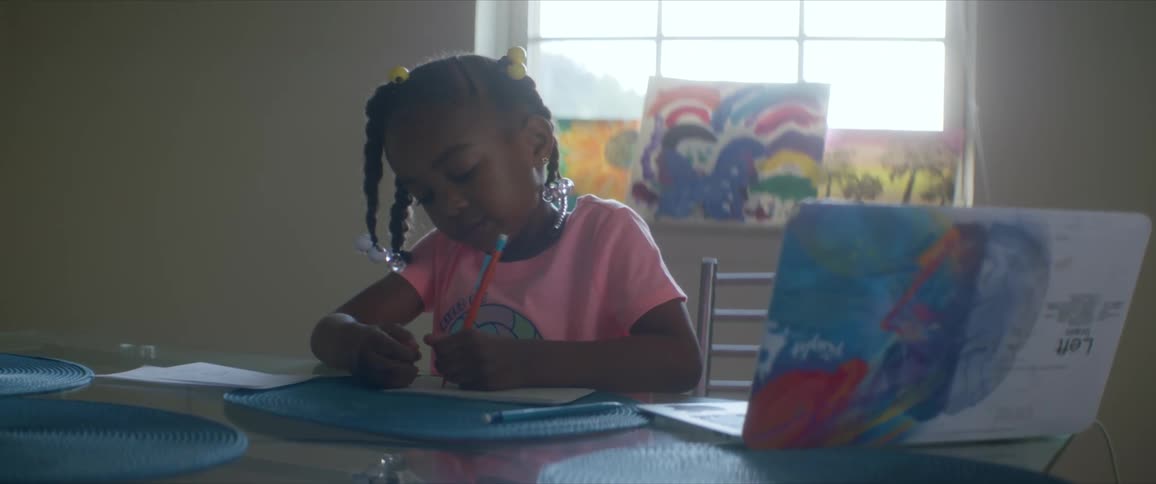 A young girl sits at a table writing with a pencil and several pieces of her artwork around her