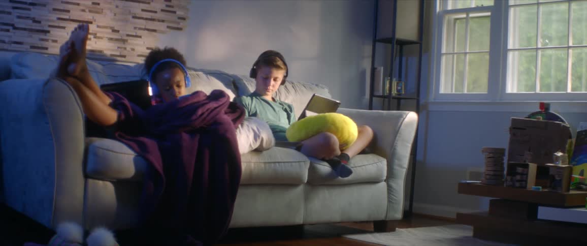 A boy and girl sit on a couch in a home and look at laptops while listening to music