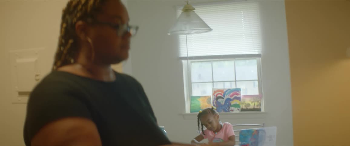 Mother stands in front of a stove in a kitchen stirring a pot while her daughter draws at the table in the dining room