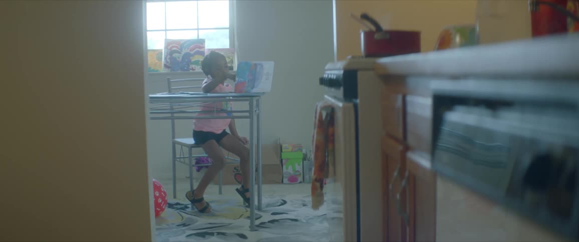 Little girl sits past a kitchen in a play room with toys and childs artwork at a table playing on a tablet