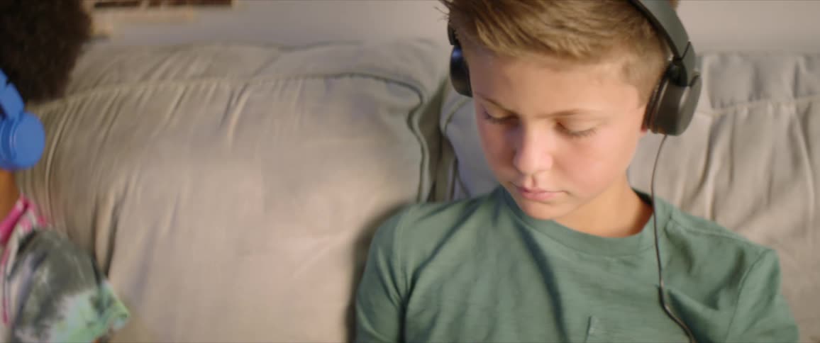 A boy and a girl sit on a couch with headphones on both watching their own devices