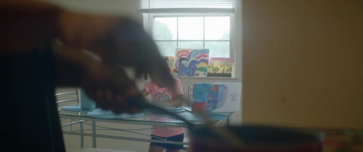 A young girl at home does school work at the table while her mom cooks