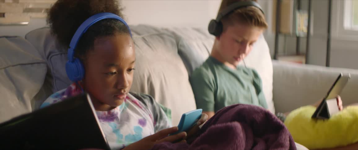 Girl wearing blue headphones and boy with black headphones and a green shirt sit on sofa and uses tablet and hand calculator in the living room at home