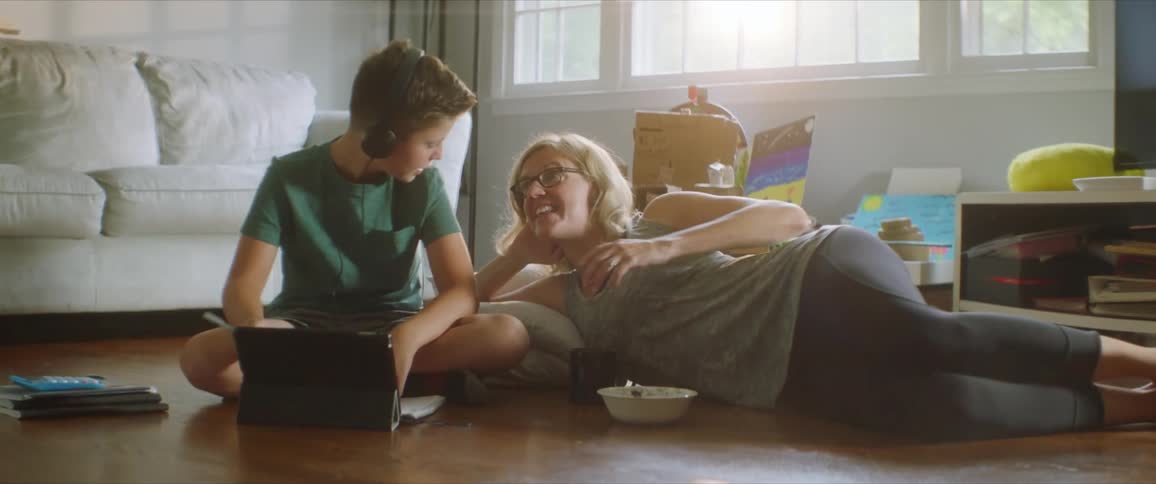 Mother laying on the floor in the living room talks to his son doing homework