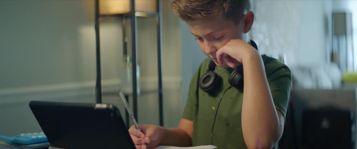 Boy with black headphones and a green shirt sits at table and writes in a notebook