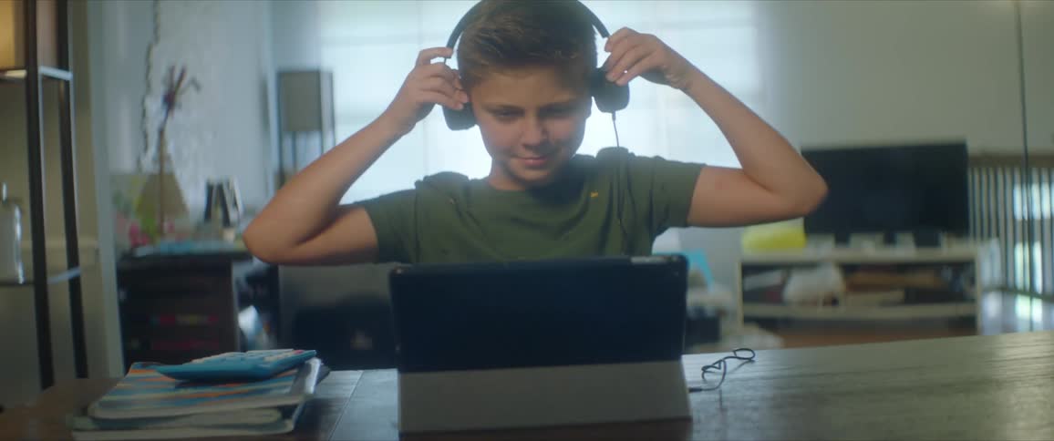 A boy smiles as he puts on his headphones and picks up his pencil to join class on his laptop