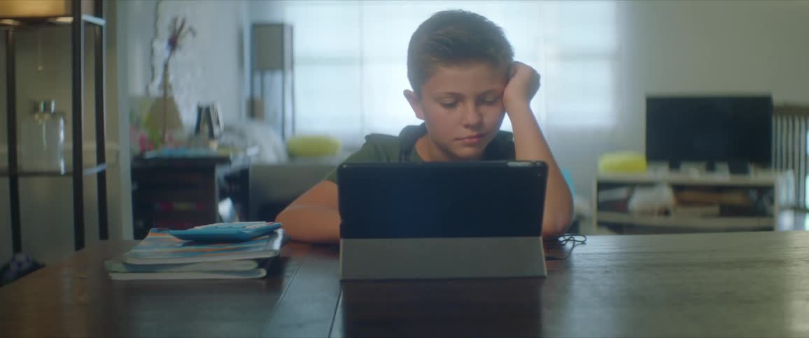 Boy with a green shirt and headphones sits at table and uses tablet in the living room at home