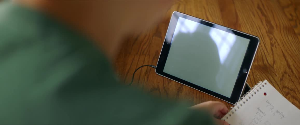 Person sits holding a notebook with writing on it in front of a tablet on the floor