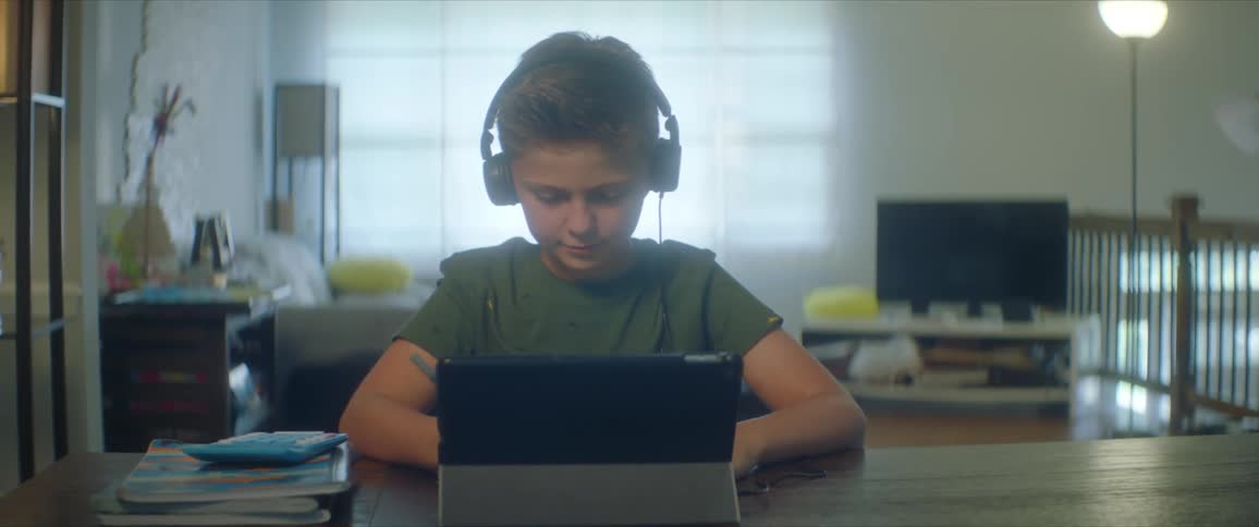 Boy with a green shirt and headphones sits at table and uses tablet in the living room at home