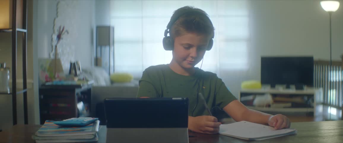 A boy with headphones sitting at a table writing in a notebook with a tablet in front of him