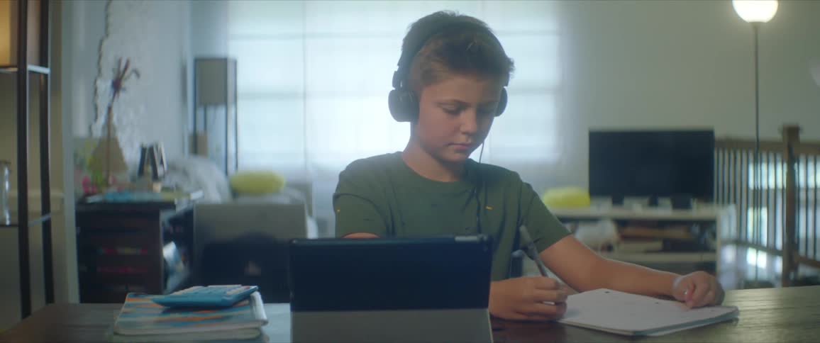 Boy with a green shirt and headphones sits at table writing in notebook and uses tablet in the living room at home