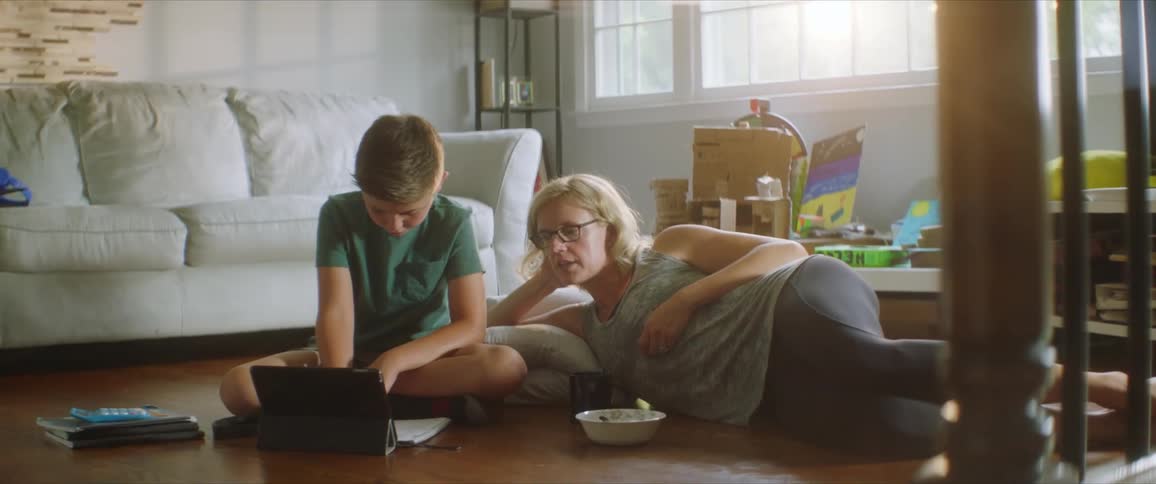 Mother lies down on floor next to son as he does school work viewing tablet