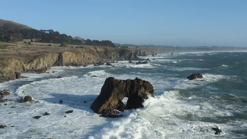 Ocean waves splash against rock formation as they roll in on a clear day