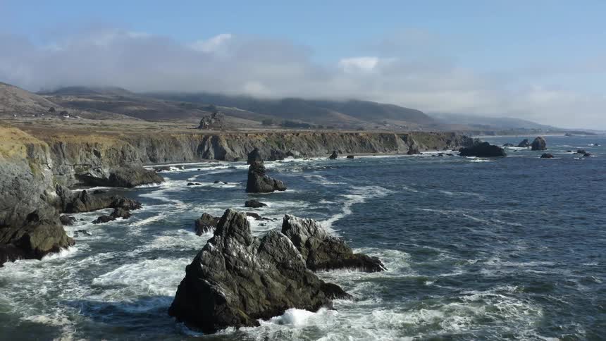 Formations of rock stick out of the ocean below the cliffs of a landmass during the day