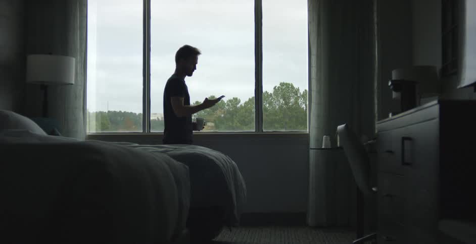 Man looking out of a window at daytime in a bedroom hanging up a phone call and walking away
