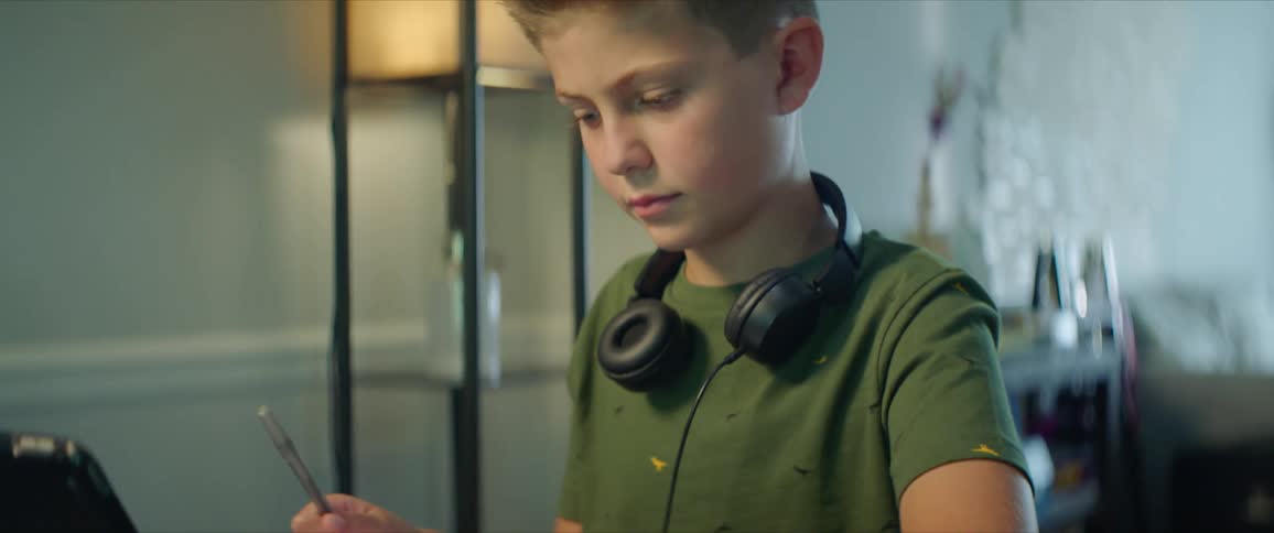 A young boy sits at a tablet with a tablet and writing in a notebook