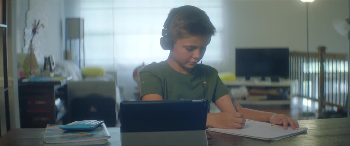 Boy with black headphones and a green shirt sitting at table writes in a notebook
