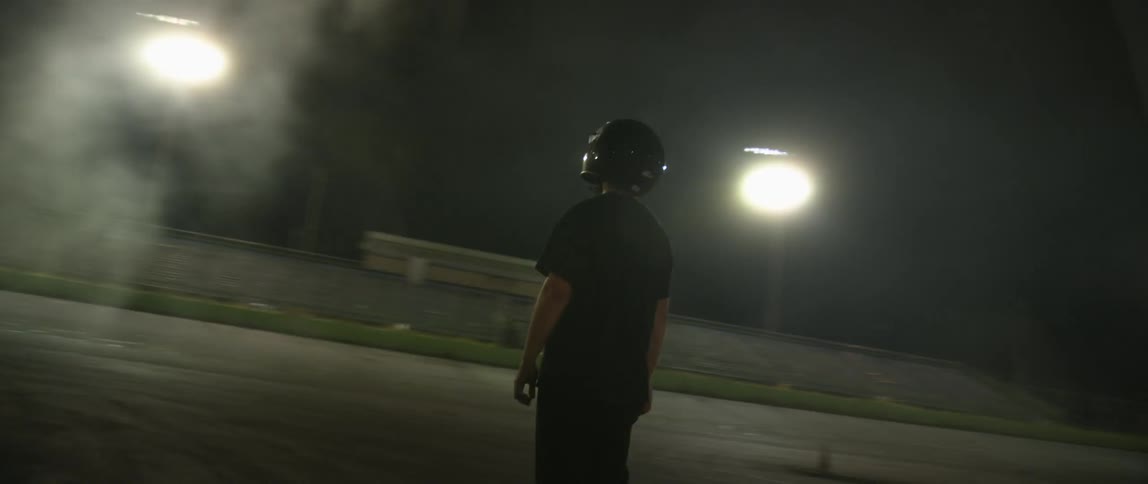 A teenager covers his face with a helmet standing in a stadium at night as the camera moves around him.