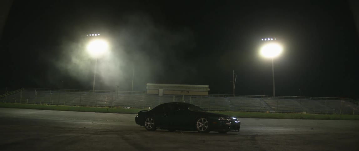 A car with its headlights being turned off with bleachers and spotlights in the background