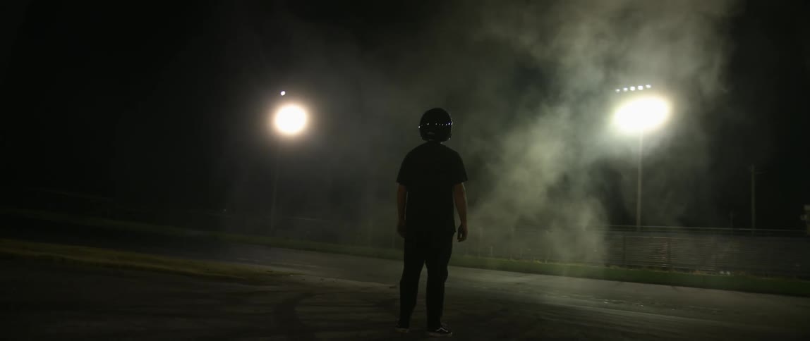 Man wearing racing helmet stands on track near smoke cloud illuminated by bright lights at night