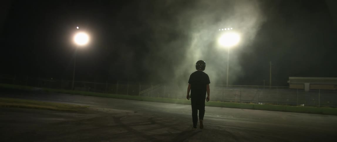 A person wearing a racing helmet walks across a smoky field next to bleachers and a track