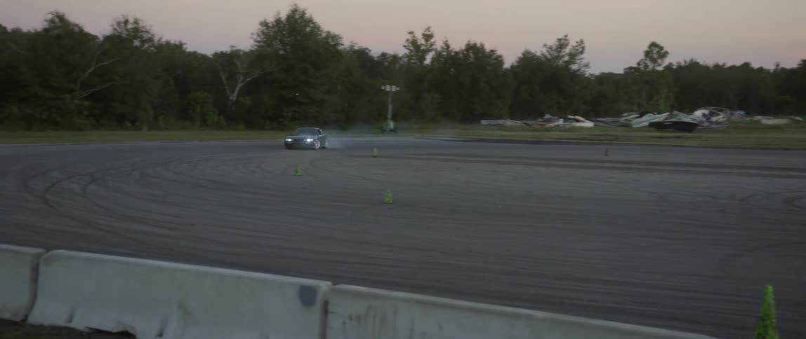 Men stand watching a car speed around an asphalt lot burning rubber as it slides through turns