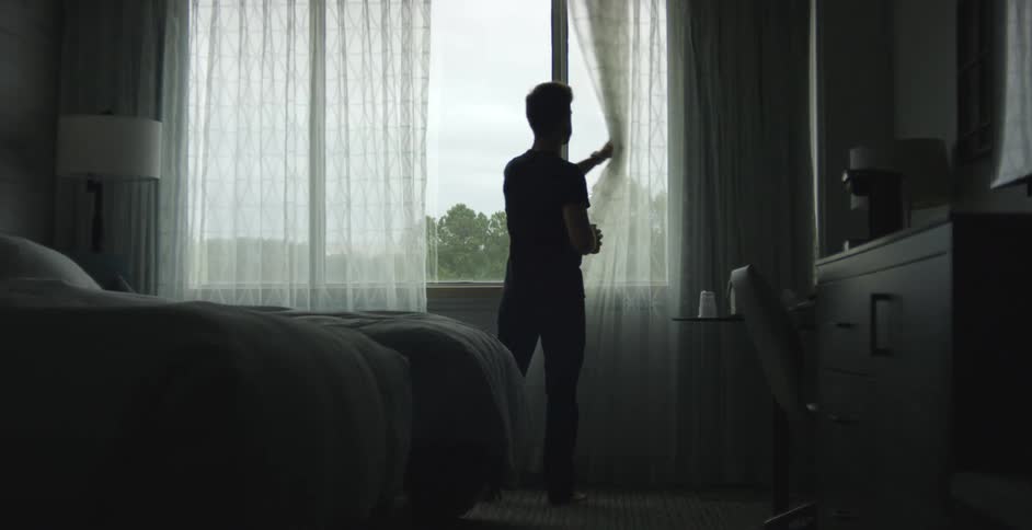 A man in a hotel picks up a cup of coffee and walks to the window and opens the drapes and drinks coffee while looking outside