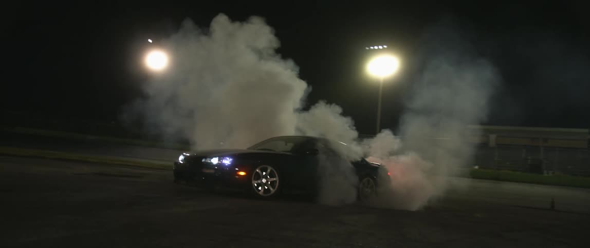 A car on the dirt infield of a stadium spins its rear tires causing smoke and exhaust to billow at night