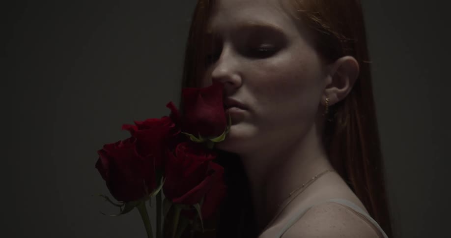 A young woman stands in a dark room while holding roses against her face
