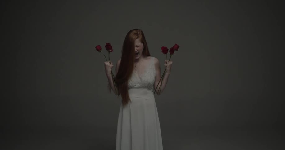 A woman stands in a room in a dress holding red roses and screaming