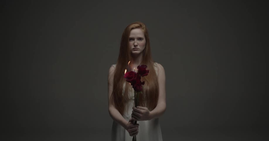 Woman holding a set of red roses on fire staring blankly ahead