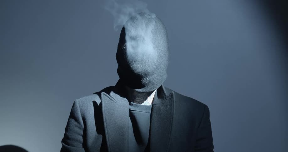 A man dressed in all black blows smoke out of a knit hat pulled down over his face and to his neck