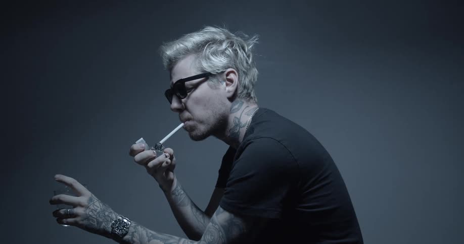 A man with tattoos wearing sunglasses and holding a glass sits and lights a cigarette in his mouth