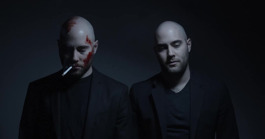 A man with blood on his face and a cigarette hanging out of his mouth stands beside his identical twin